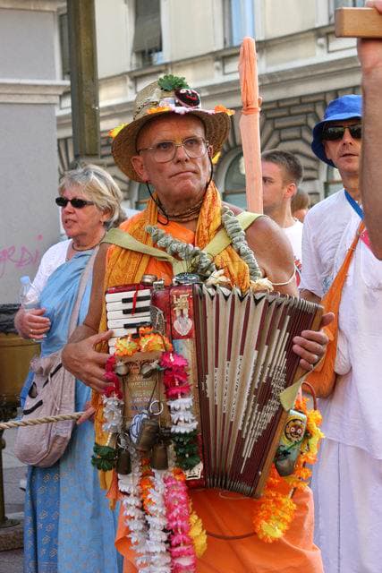 Mahavishnu Swami in a colourful hat, playing an accordion garlanded with flowers at a street kirtan
