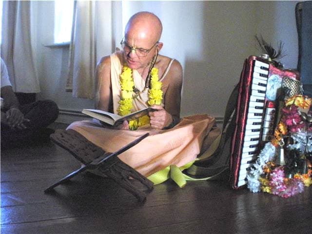 Mahavishnu Swami seated reading from a book on a stand, a decorated harmonium beside him