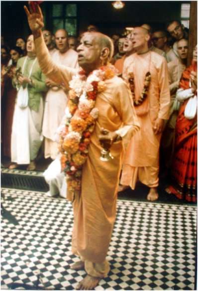 Srila Prabhupada in saffron with a flower garland, arm raised, leading kirtan among his disciples